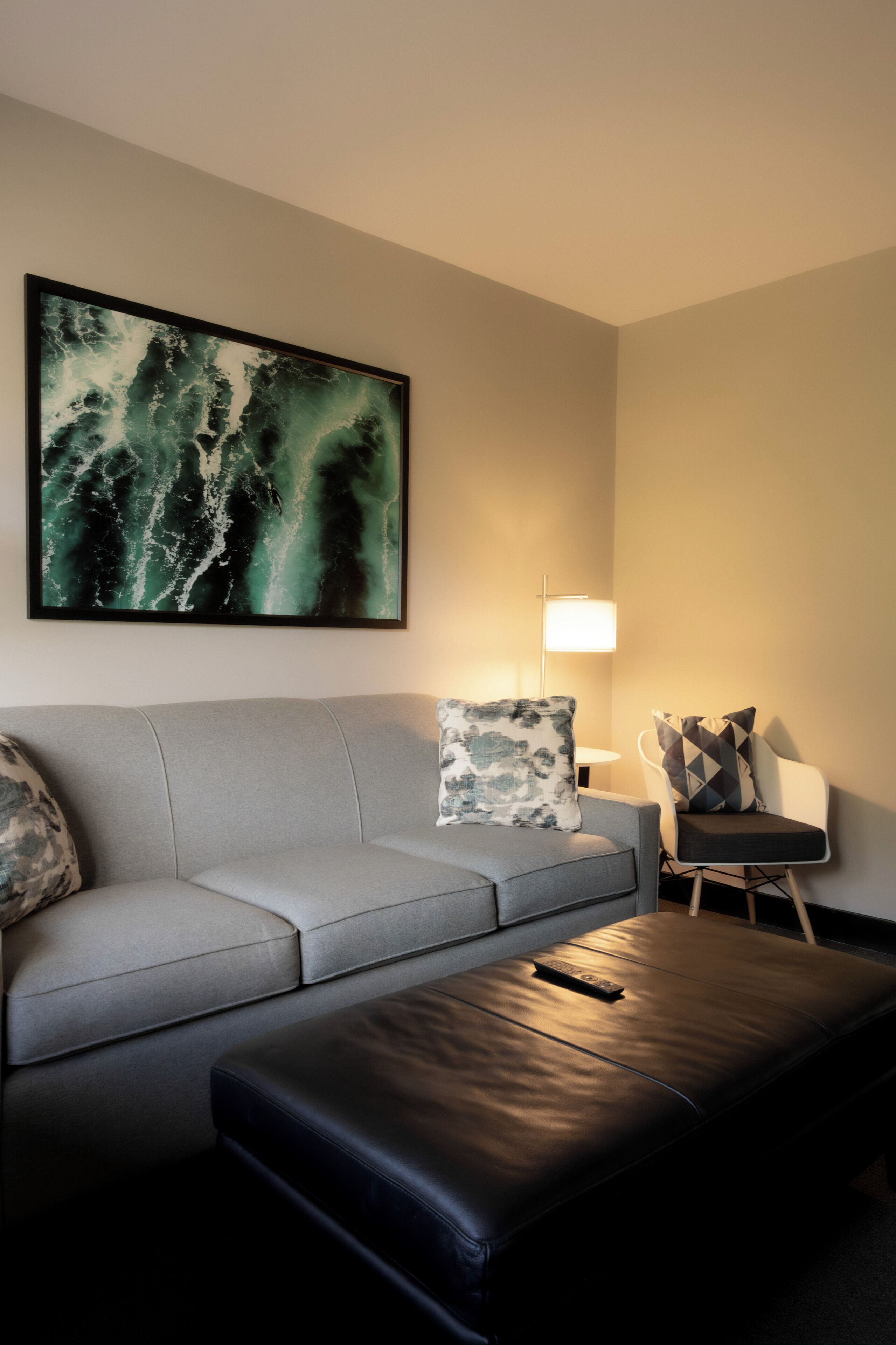 A modern living room with a grey sofa, a black ottoman, a small round table with a lamp, and a framed abstract painting on the wall.