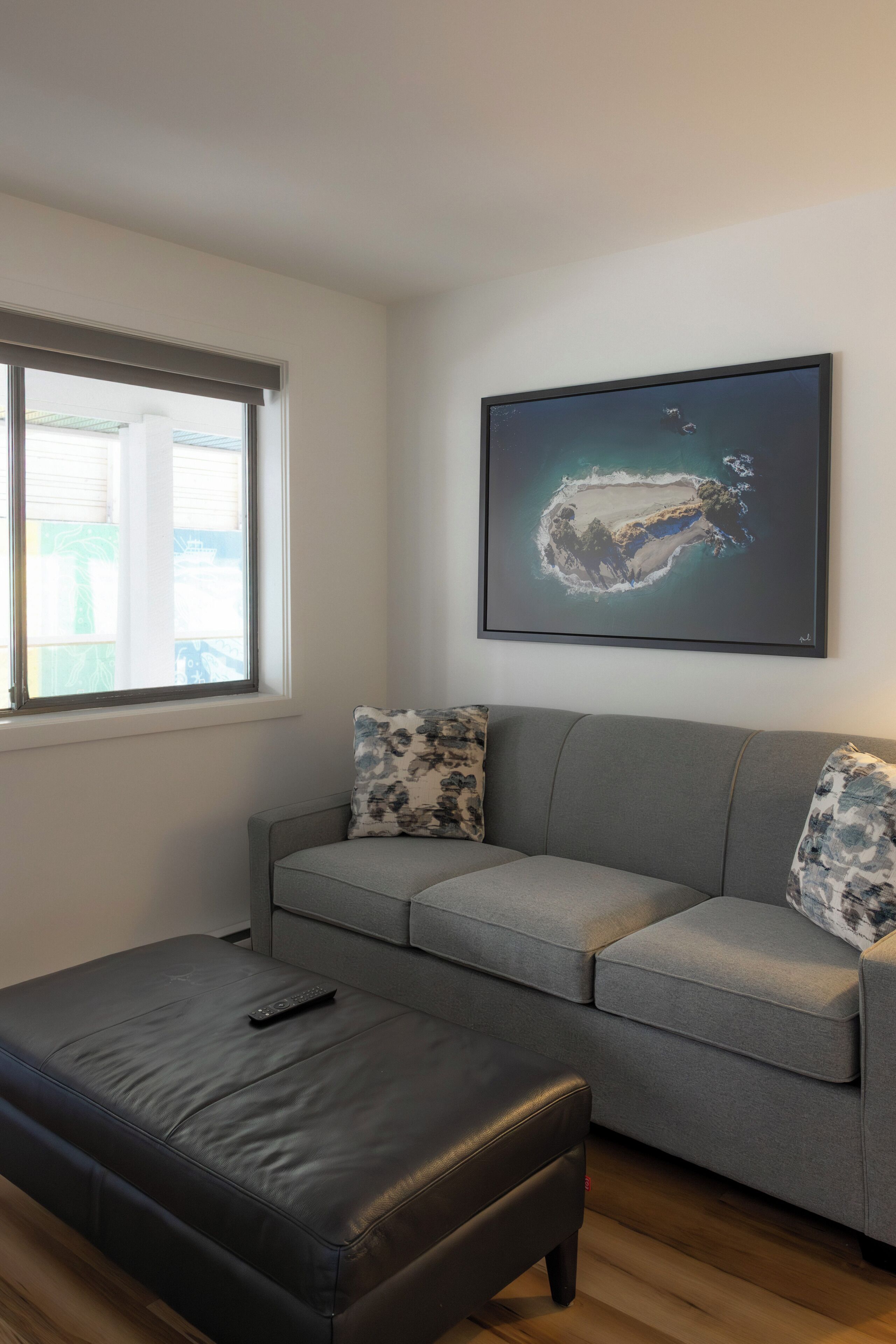 A modern living room with a grey sofa, a black ottoman, and a framed artwork on the wall.