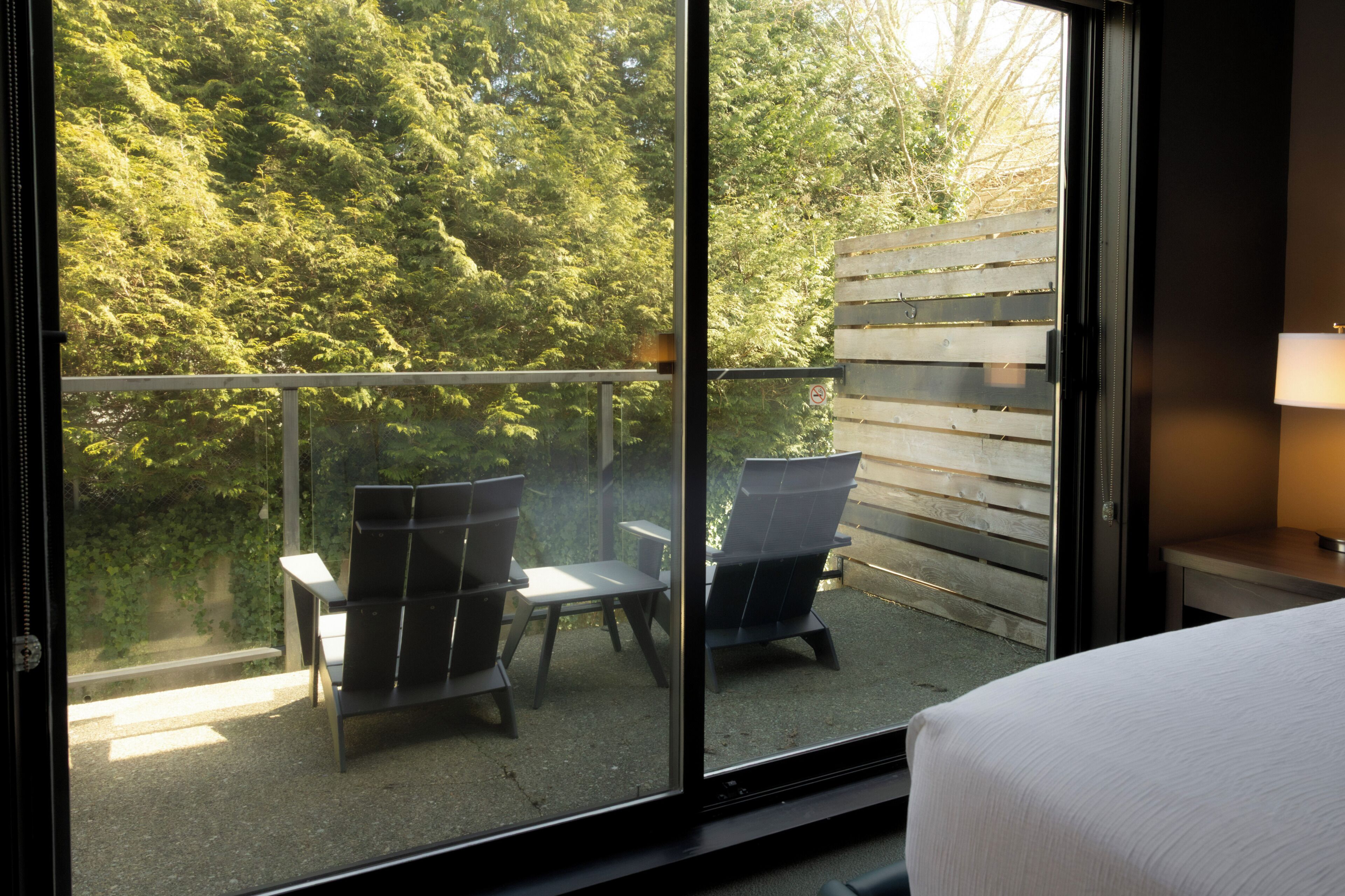 A balcony with two chairs and a table, a bed, and a view of a wooden fence and trees.