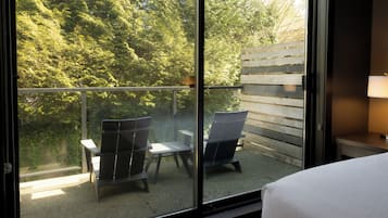 Ein Balkon mit zwei Stühlen, einem Tisch, einem Bett und Blick auf einen Holzzaun und Bäume.