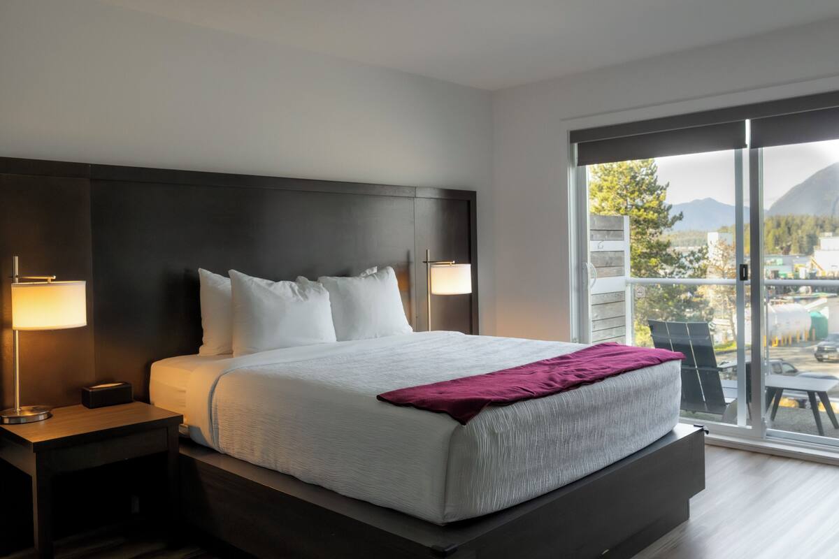 Een moderne hotelkamer met een groot bed, een nachtkastje, een lamp en uitzicht op het landschap door een groot raam.