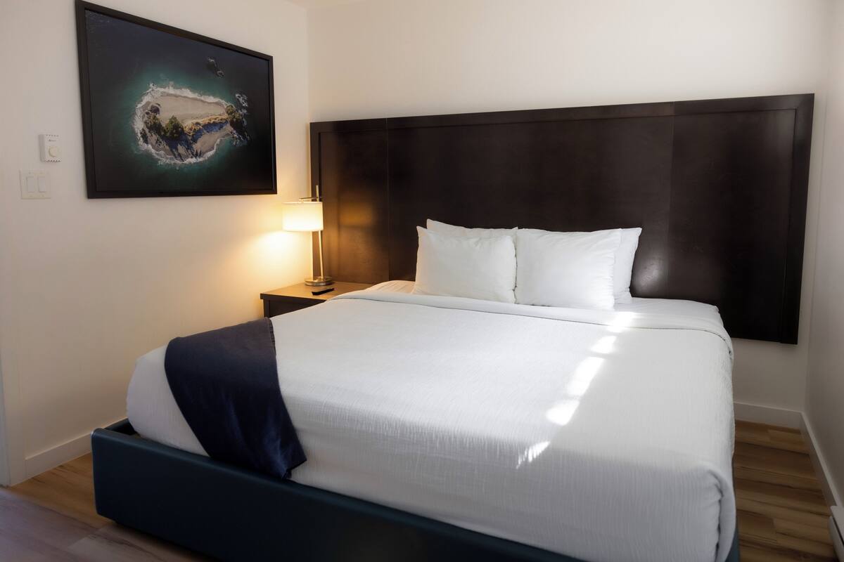 Een hotelkamer met een groot bed, een donker hoofdeinde, een nachtkastje met een lamp en een lijst met een afbeelding aan de muur.