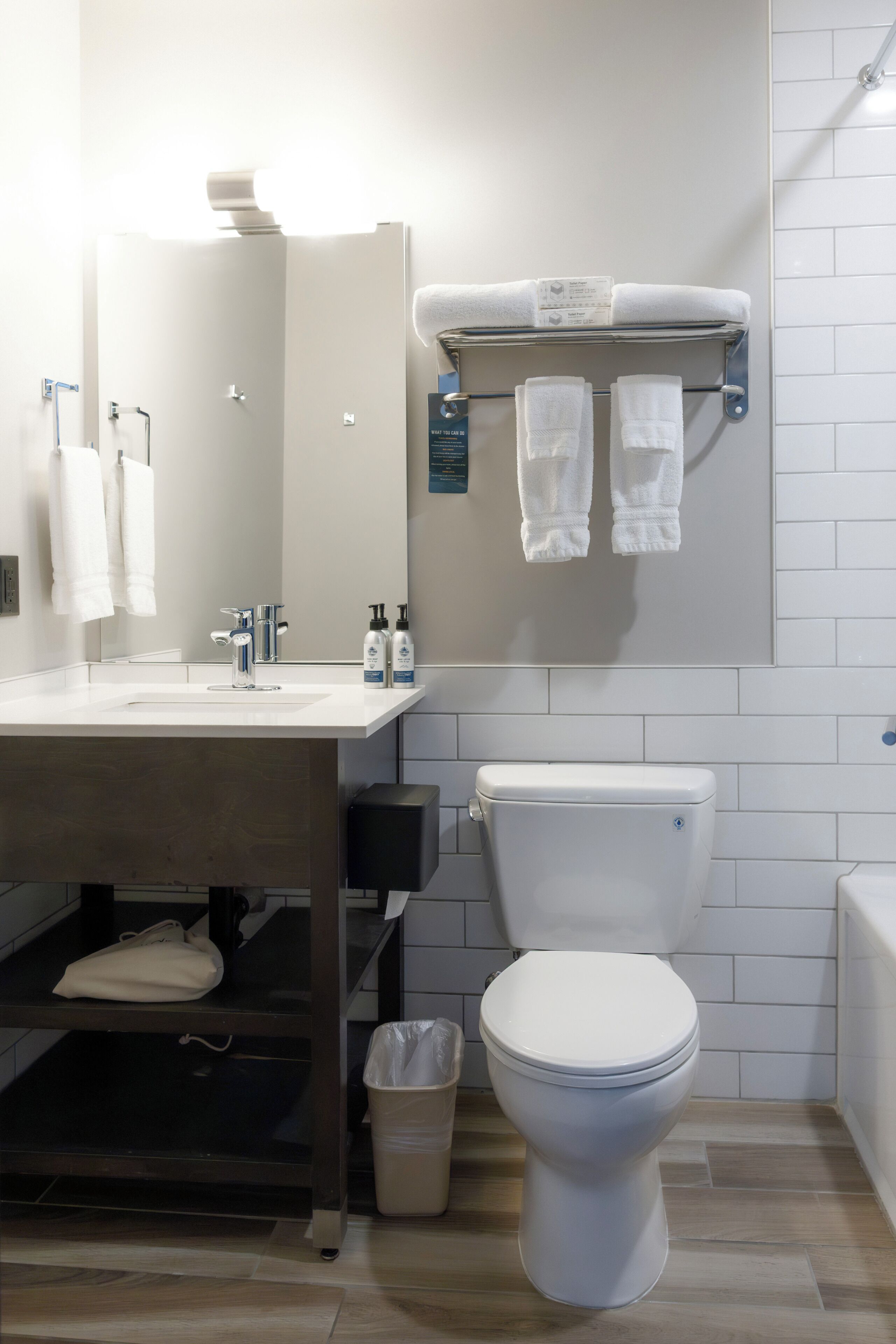 A modern bathroom with a white sink, mirror, and toilet. There are towels hanging on a rack and a trash bin.