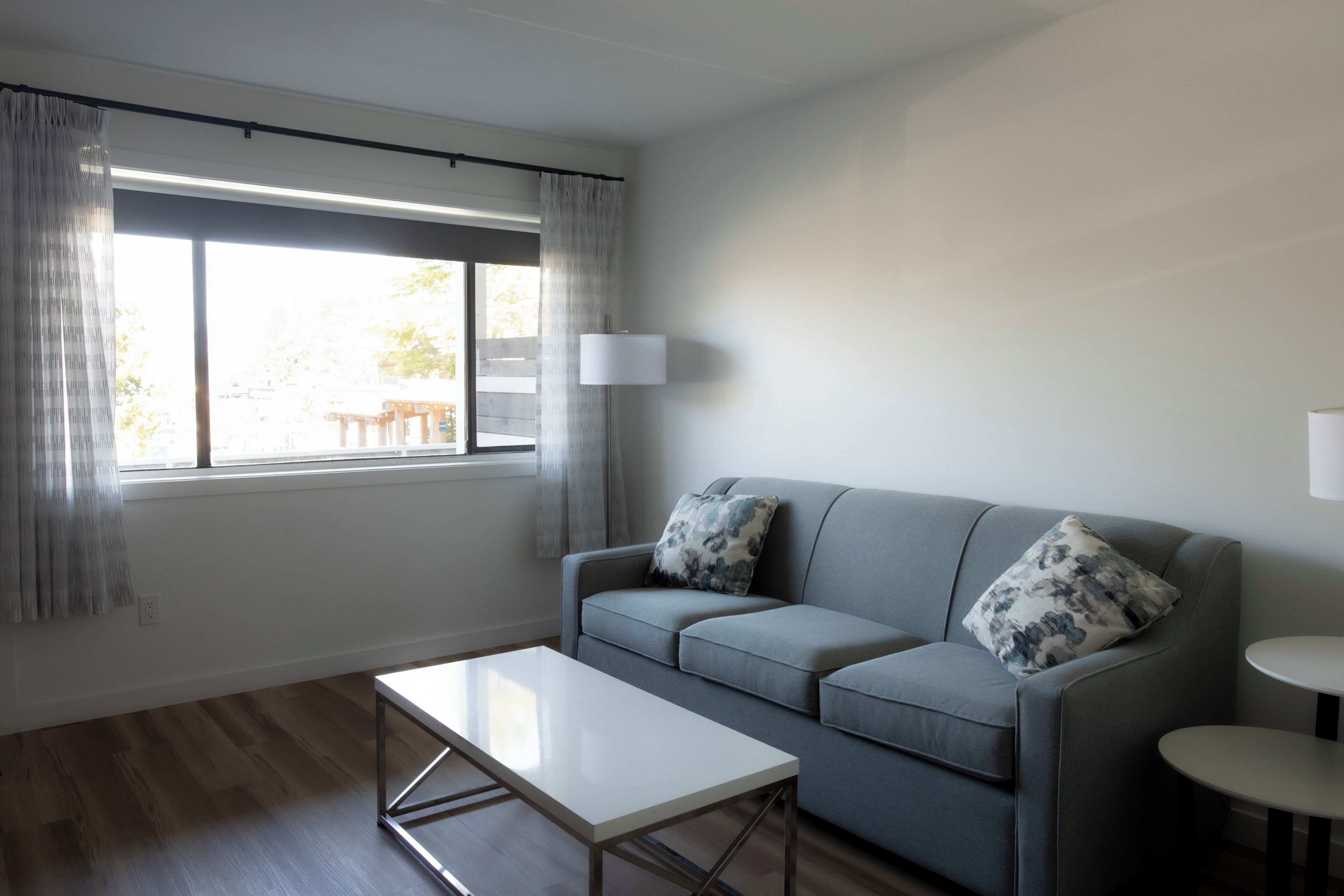Ett modernt vardagsrum med en grå soffa, två kuddar, ett vitt soffbord, en golvlampa och ett fönster med gardiner.