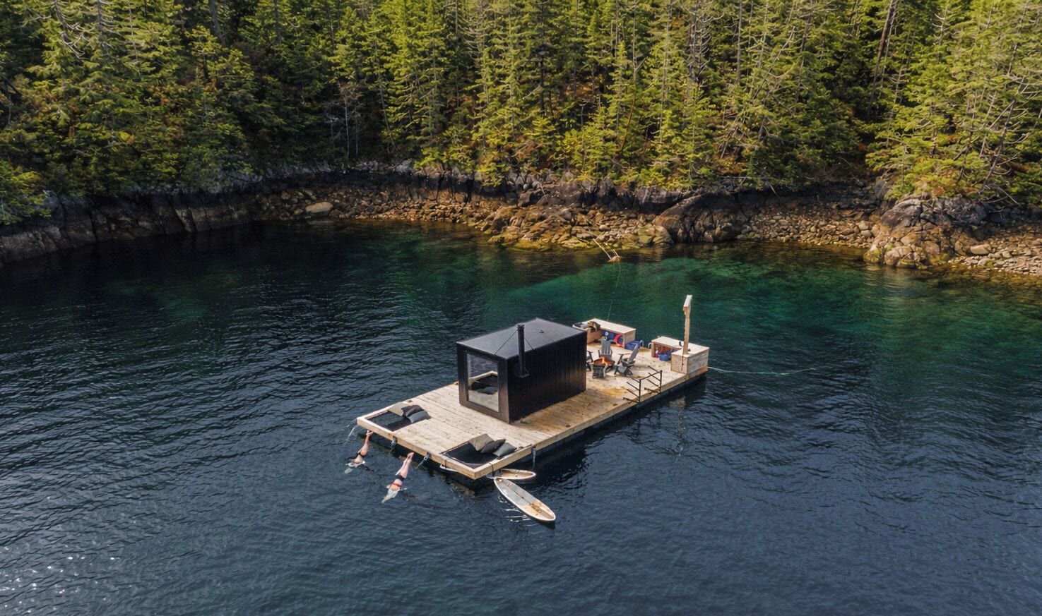 A floating structure with a small deck, seating, and a covered area, surrounded by clear water and dense forest.