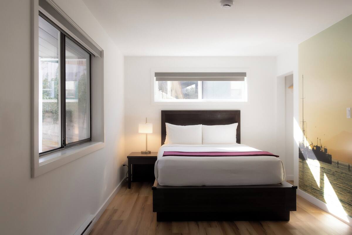 Een slaapkamer met een bed, een nachtkastje, een raam en een kunstwerk aan de muur.