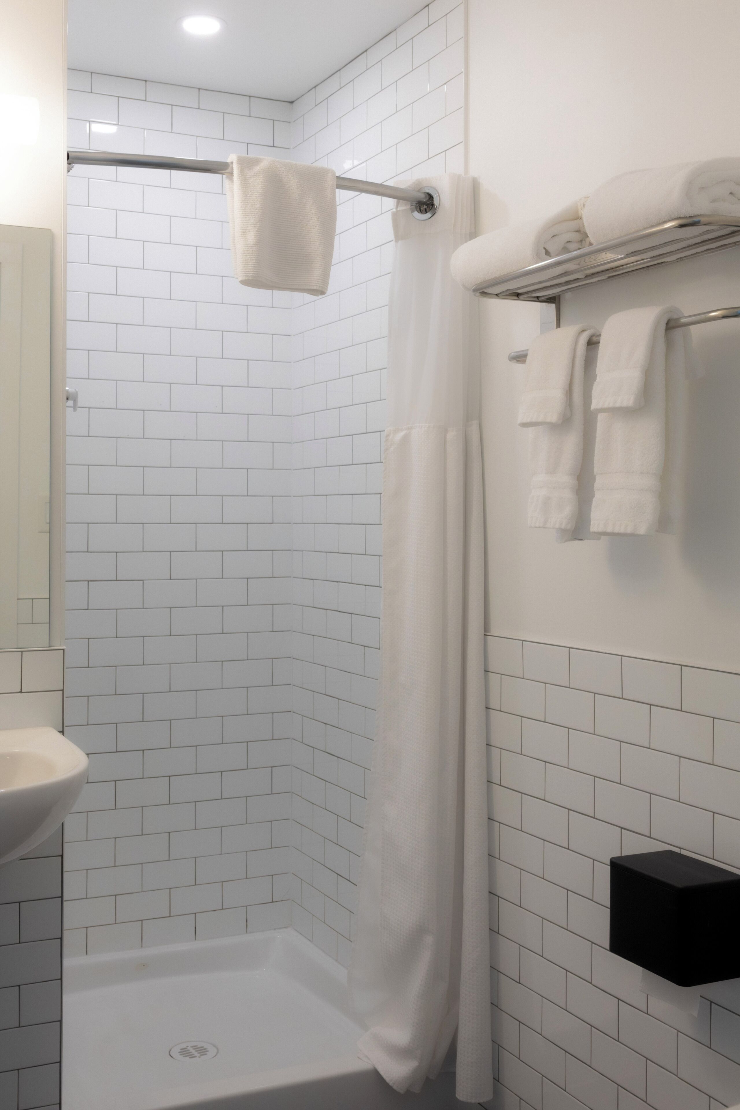 Un baño con paredes de azulejos blancos, una ducha con cortina blanca, un lavabo y un perchero con toallas.
