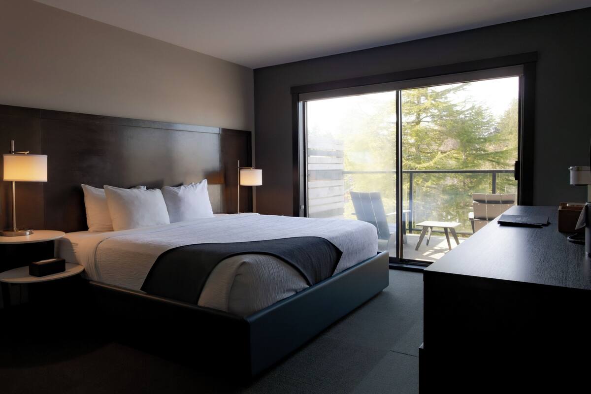 Een moderne hotelkamer met een groot bed, twee nachtkastjes met lampen, een bureau en uitzicht door een groot raam.
