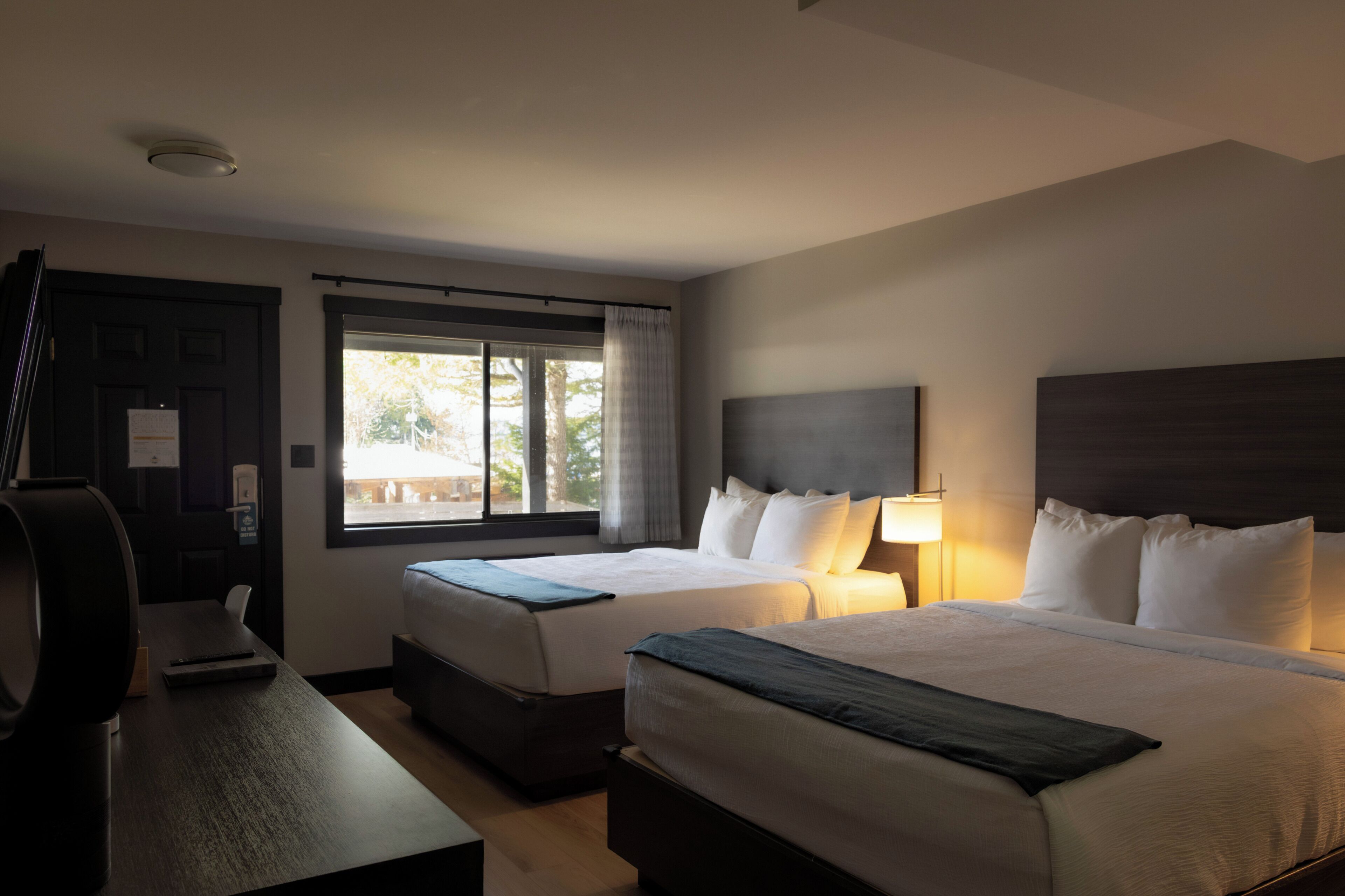 A hotel room with two beds, a desk, a window with curtains, and a headboard with a lamp.