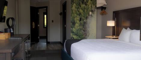 A hotel room with a bed, a desk, a trash bin, a lamp, and a wall with a nature-themed mural.
