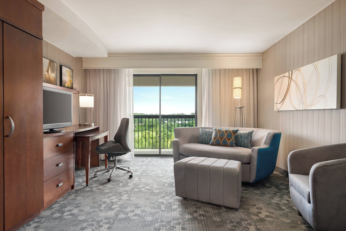 A hotel room with a desk, television, sofa, ottoman, and a view of greenery through the window.