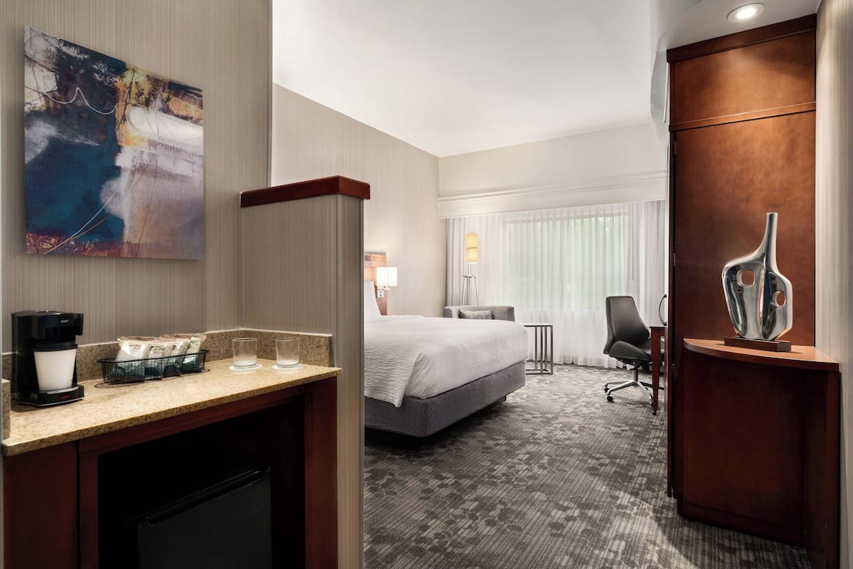 A hotel room with a bed, a desk with a coffee maker, a chair, and a large abstract painting on the wall.