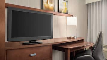 Habitación de hotel con una televisión de pantalla plana grande sobre un escritorio de madera, una lámpara, una silla y un cajón pequeño.