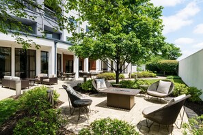 Un espacio al aire libre moderno con mobiliario de mimbre y una mesa central, rodeado de follaje y la fachada de un edificio.