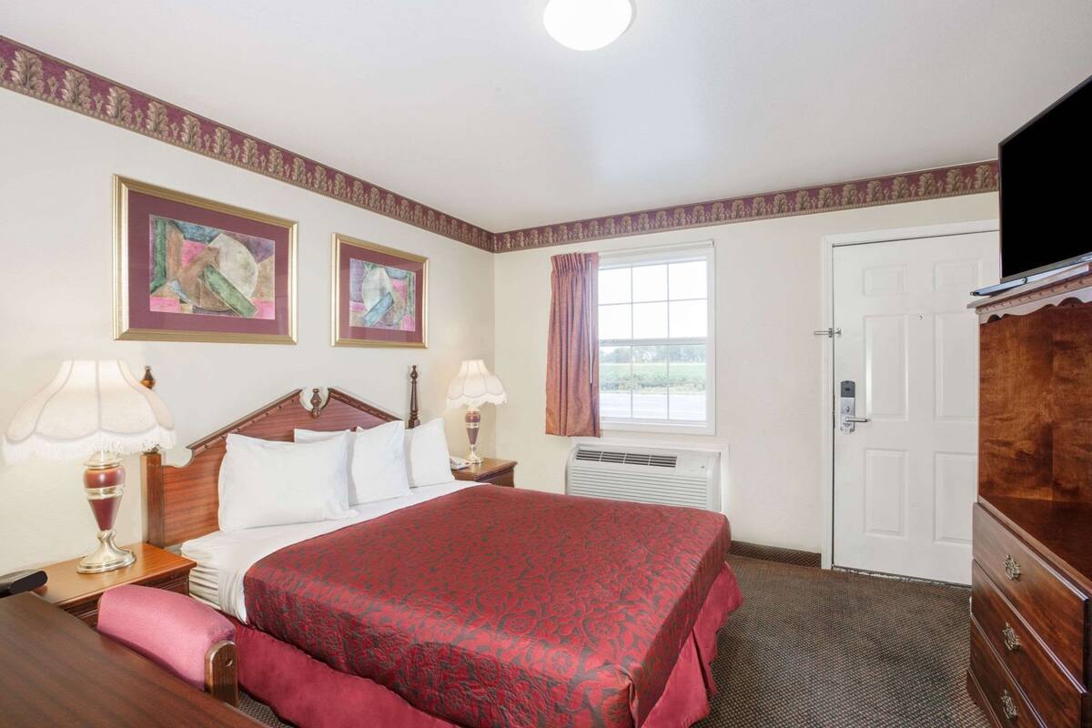 A hotel room with a bed, two bedside tables with lamps, a TV, a window with curtains, and two framed pictures on the wall.