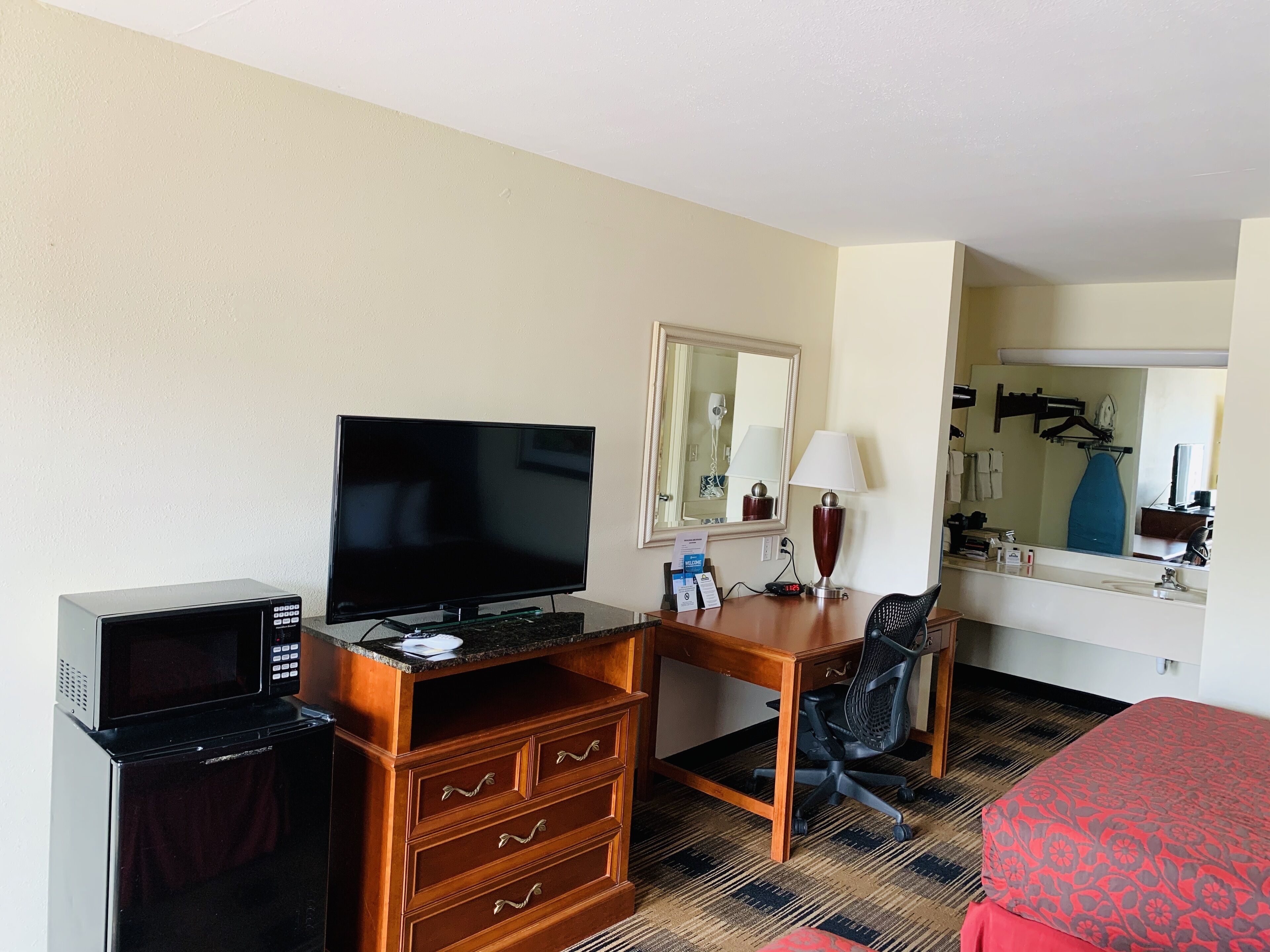A hotel room with a flat-screen TV, a wooden desk with drawers, a chair, a microwave, a lamp, and a bed with a patterned cover.