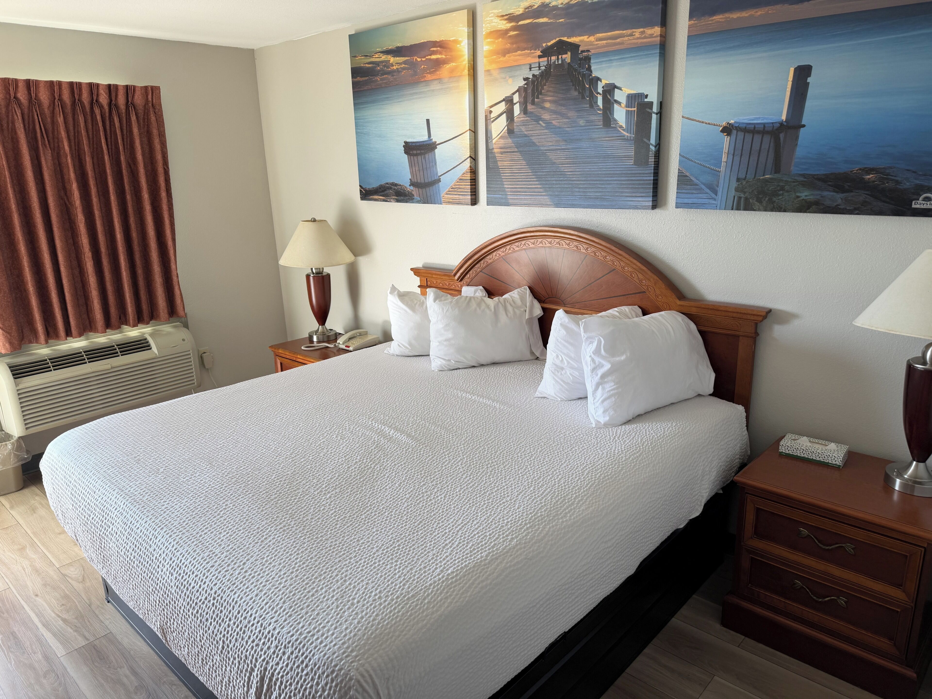 A hotel room with a large bed, wooden headboard, white bedding, two bedside lamps, a telephone, and a wall-mounted air conditioner.