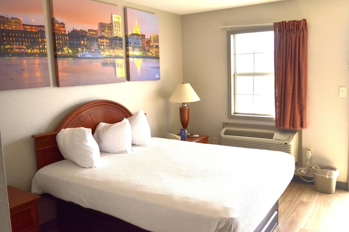 A hotel room with a bed, a nightstand, a lamp, a window with curtains, and artwork on the wall.