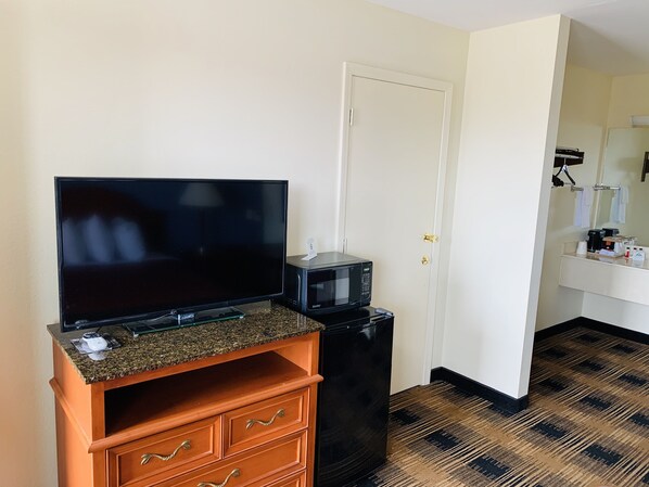 A hotel room with a flat-screen TV on a wooden cabinet, a microwave, and a door leading to a bathroom with a sink and mirror.