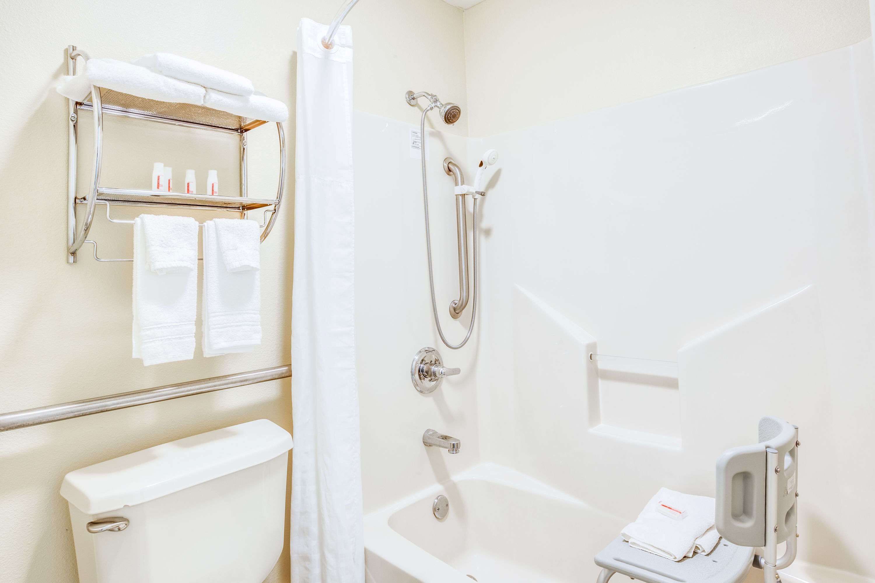 Une salle de bain comprenant des toilettes, une douche avec un pommeau de douche amovible, une baignoire équipée d’une barre d’appui et une étagère contenant des articles de toilette.