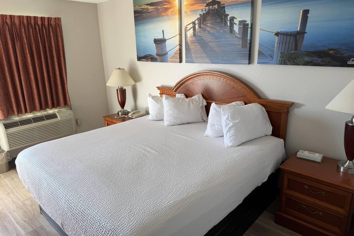 A hotel room with a large bed, a wooden headboard, a nightstand with a lamp, a wall-mounted phone, and a painting of a pier.