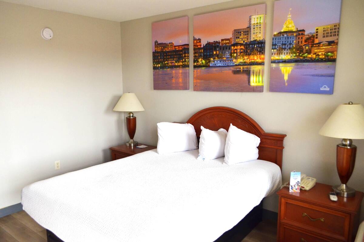 A hotel room with a large bed, two bedside tables, a nightstand, and three cityscape paintings on the wall.