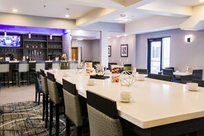 A hotel dining area with a long table, chairs, and a bar area with shelves and a television. - Holiday Inn Longview - North by IHG (Longview)