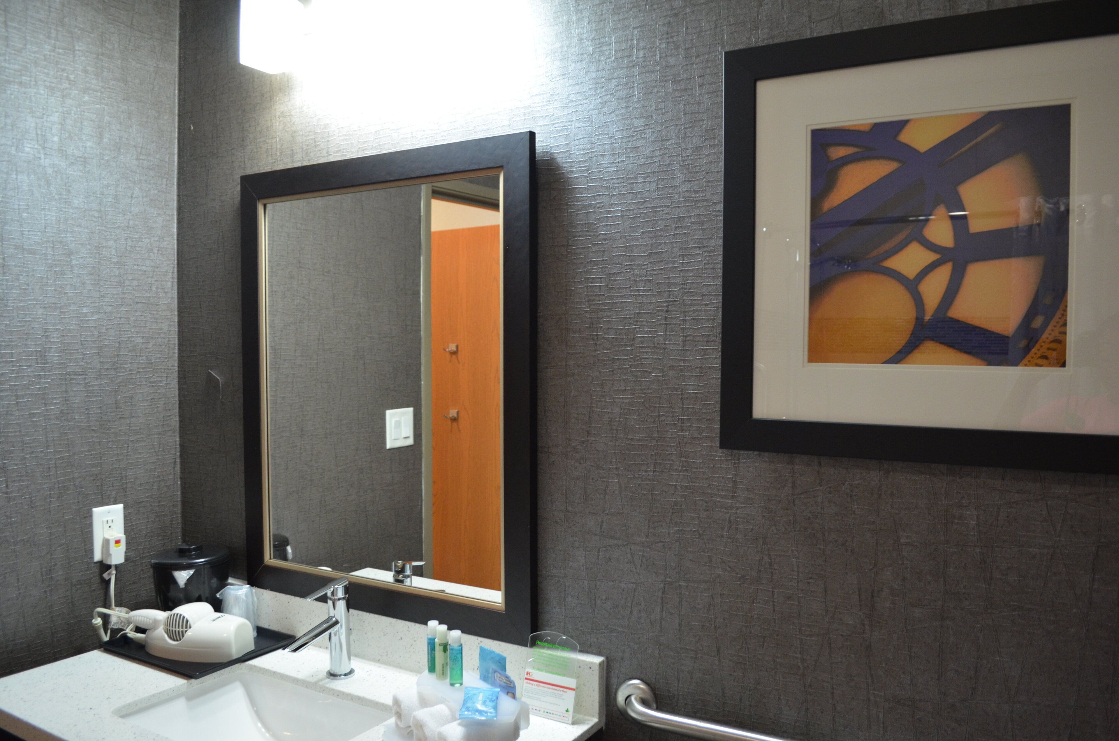 A bathroom with a large mirror, a sink, toiletries, and a framed abstract painting on the wall.