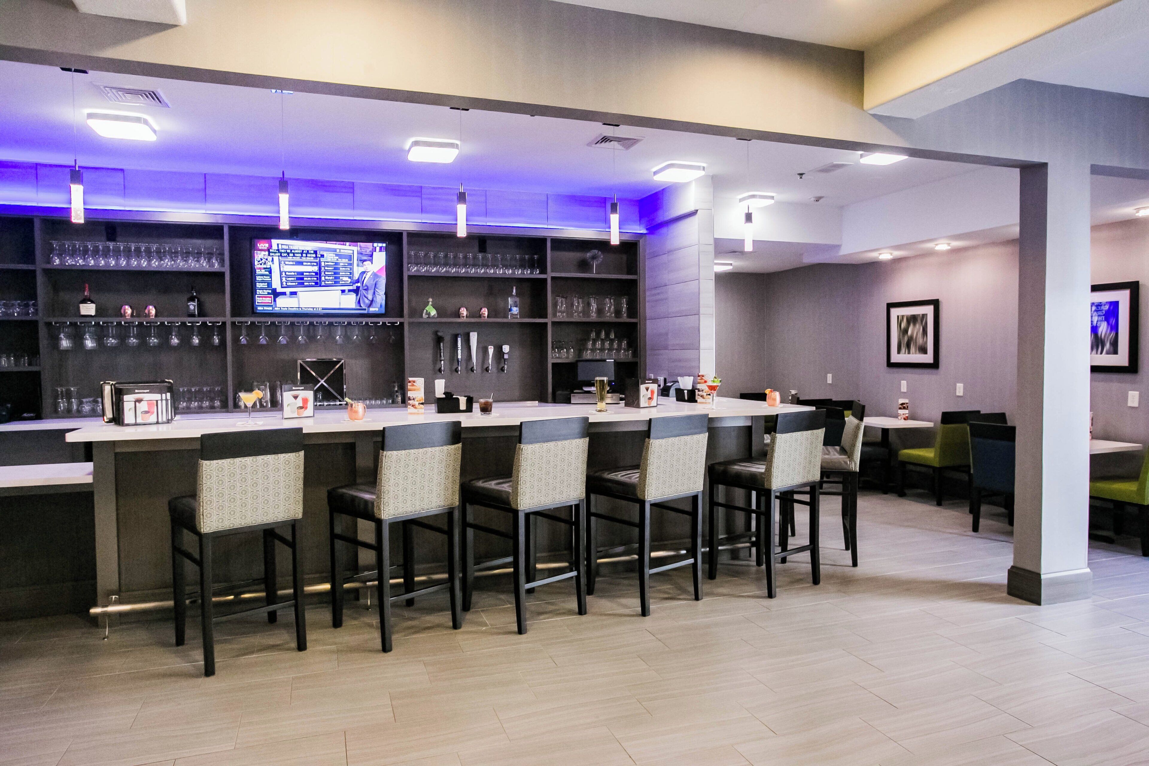 A bar area with a long counter, multiple bar stools, and a television screen displaying a menu.