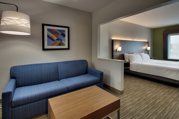 Une chambre d’hôtel avec un canapé bleu, une table basse en bois, un lit avec du linge de lit blanc, une tête de lit dotée de lampes murales et un tableau abstrait encadré accroché au mur.