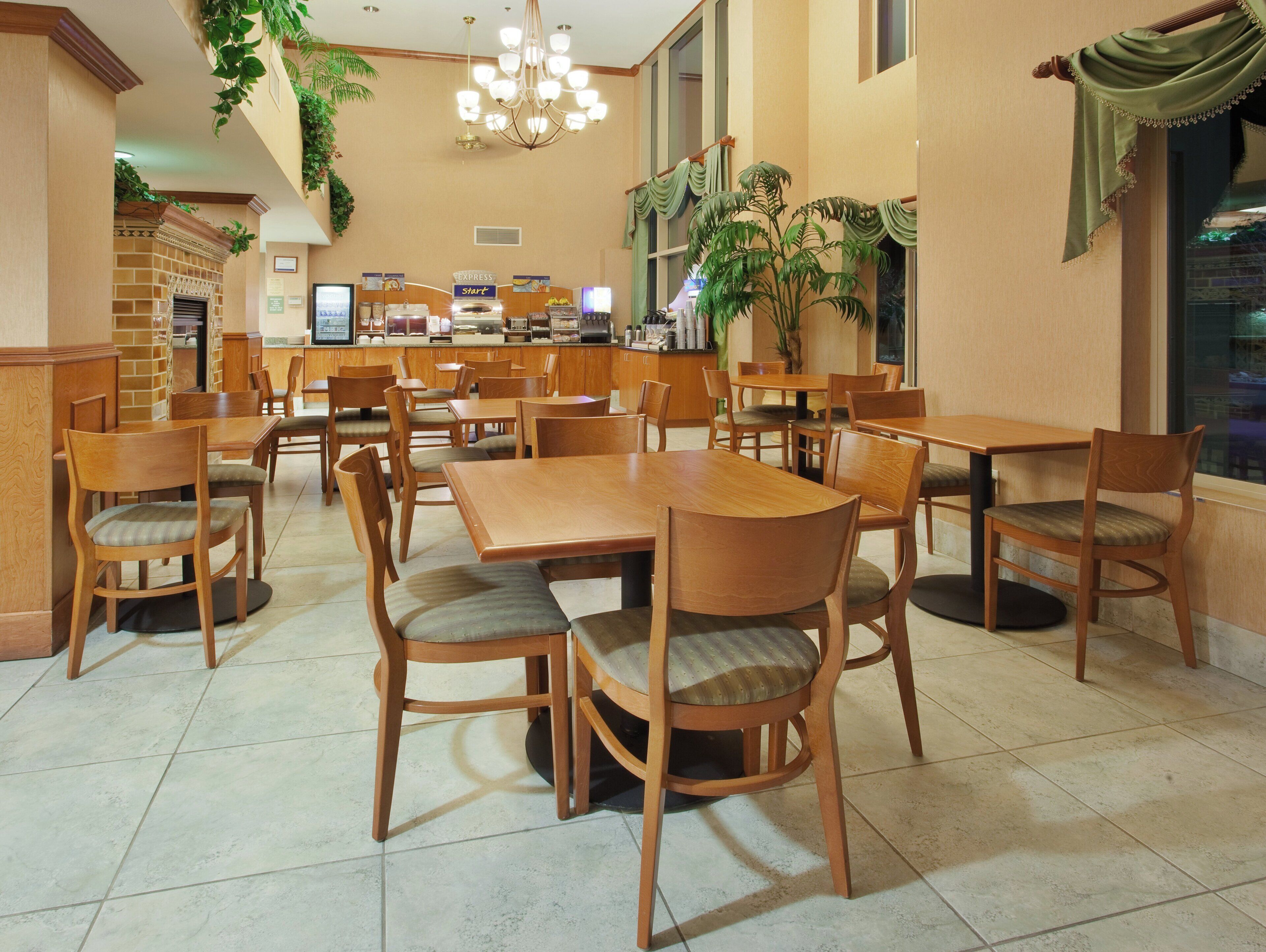 A hotel dining area with wooden tables and chairs, a chandelier, and a kitchen in the background.