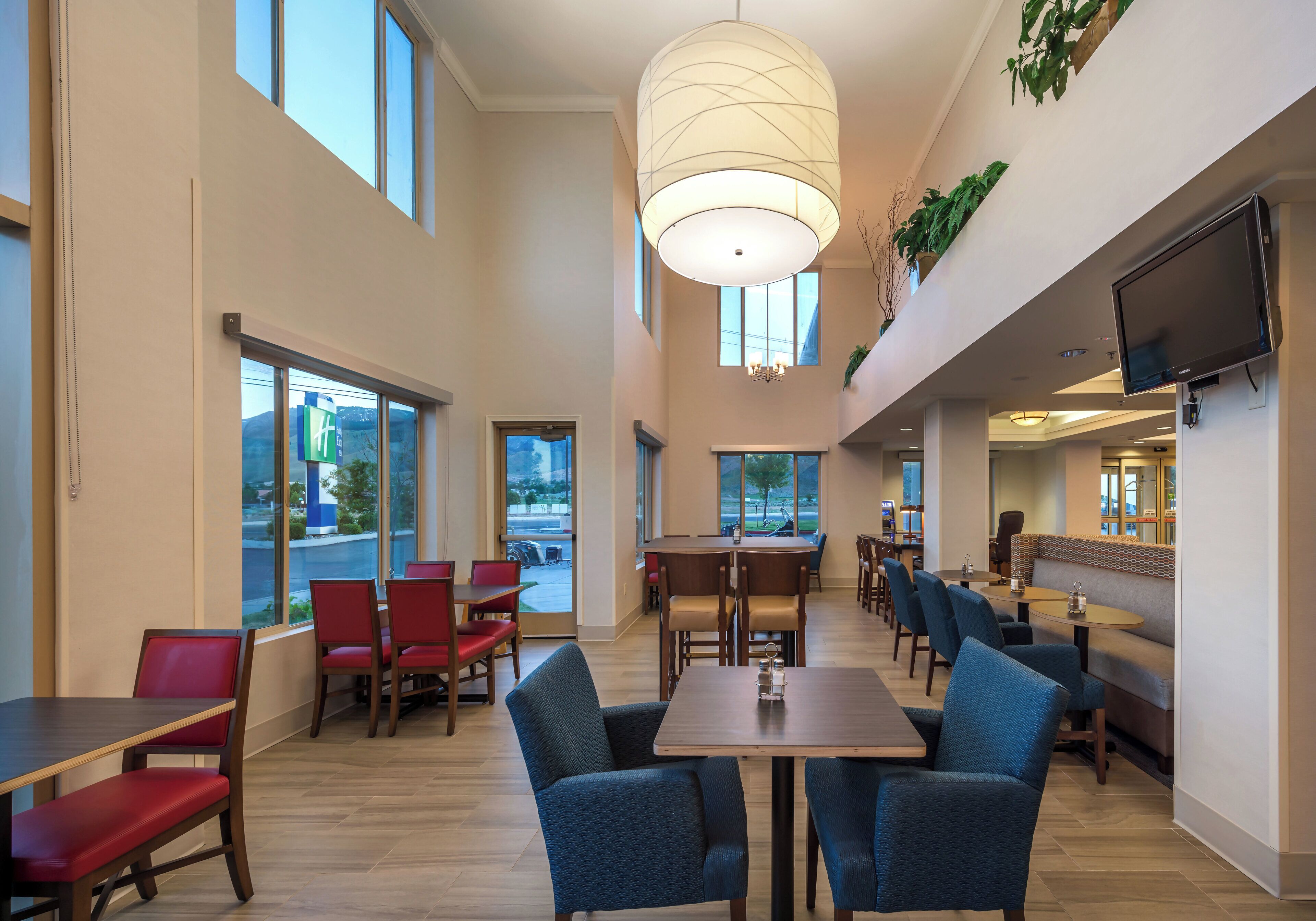A hotel dining area with multiple tables and chairs, large windows, and a television mounted on the wall.