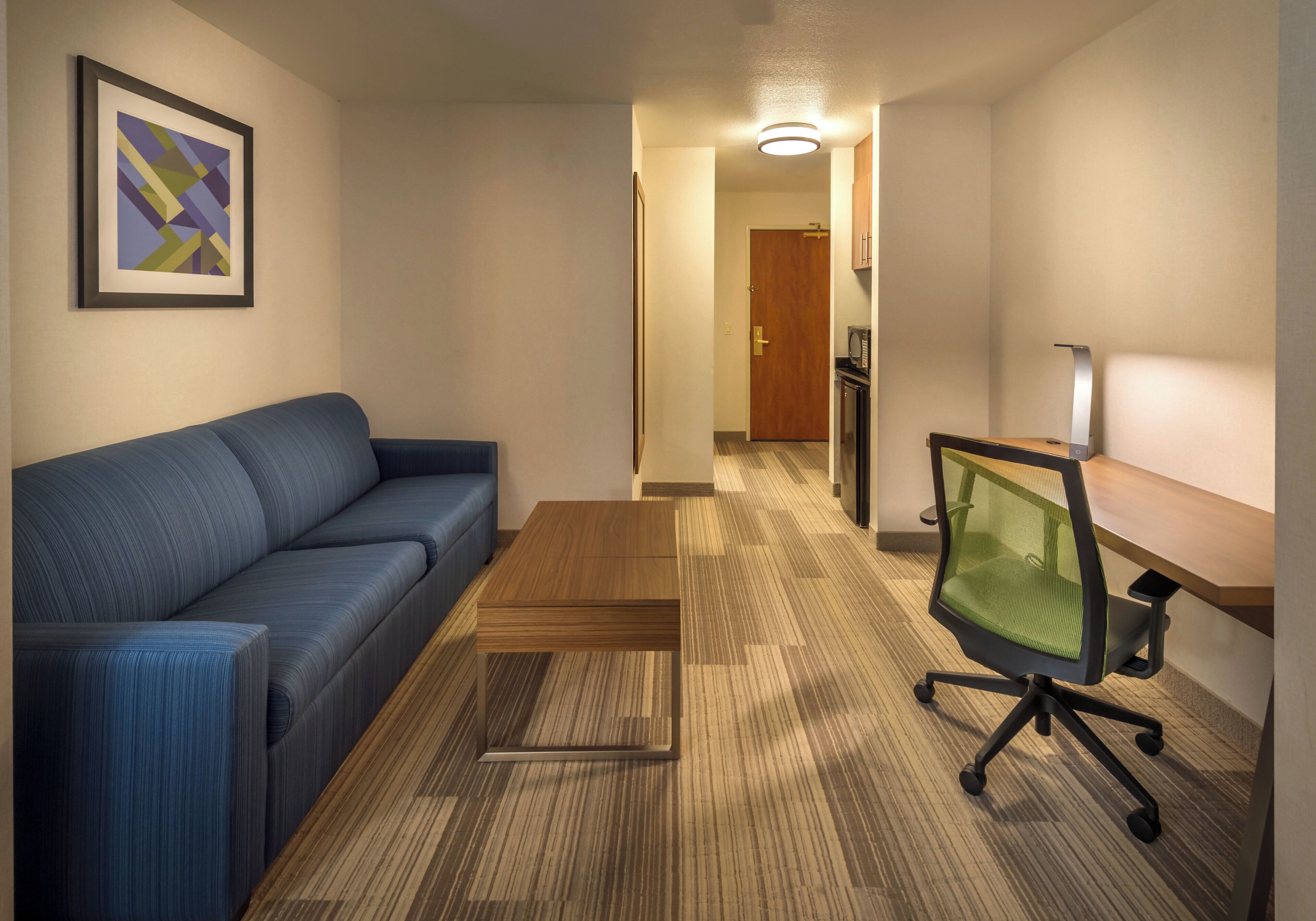 A hotel room with a blue sofa, a wooden coffee table, a green mesh office chair, a microwave, and a framed abstract artwork on the wall.