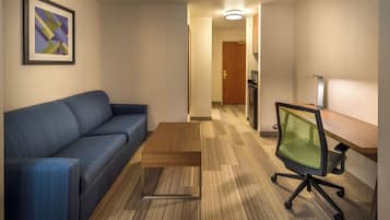 A hotel room with a blue sofa, a wooden coffee table, a green mesh office chair, a microwave, and a framed abstract artwork on the wall.