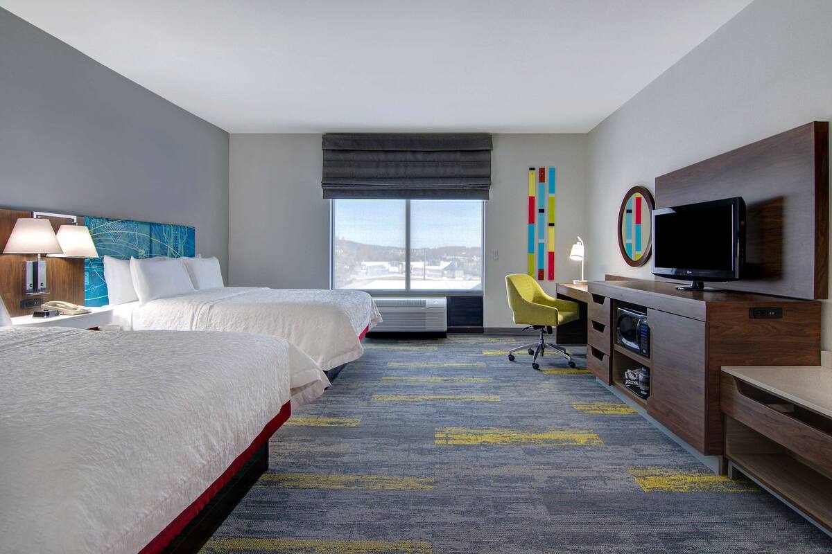 A hotel room with two beds, a desk with a TV, a chair, and a large window.