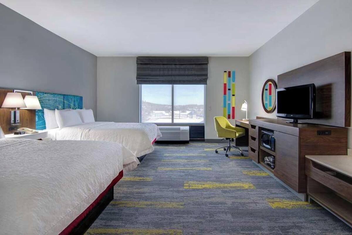 A hotel room with two beds, a desk with a TV, a chair, and a large window.