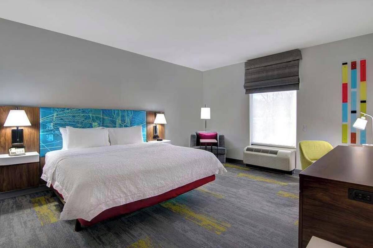 A hotel room with a bed, bedside lamps, a desk, a chair, a window with a view, and a colorful abstract wall art piece.