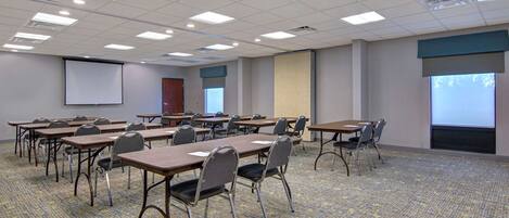 A conference room with multiple tables and chairs, a projector screen, and a carpeted floor.