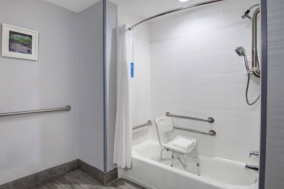 A bathroom with a walk-in shower, a white bathtub with a small chair, and a handrail.