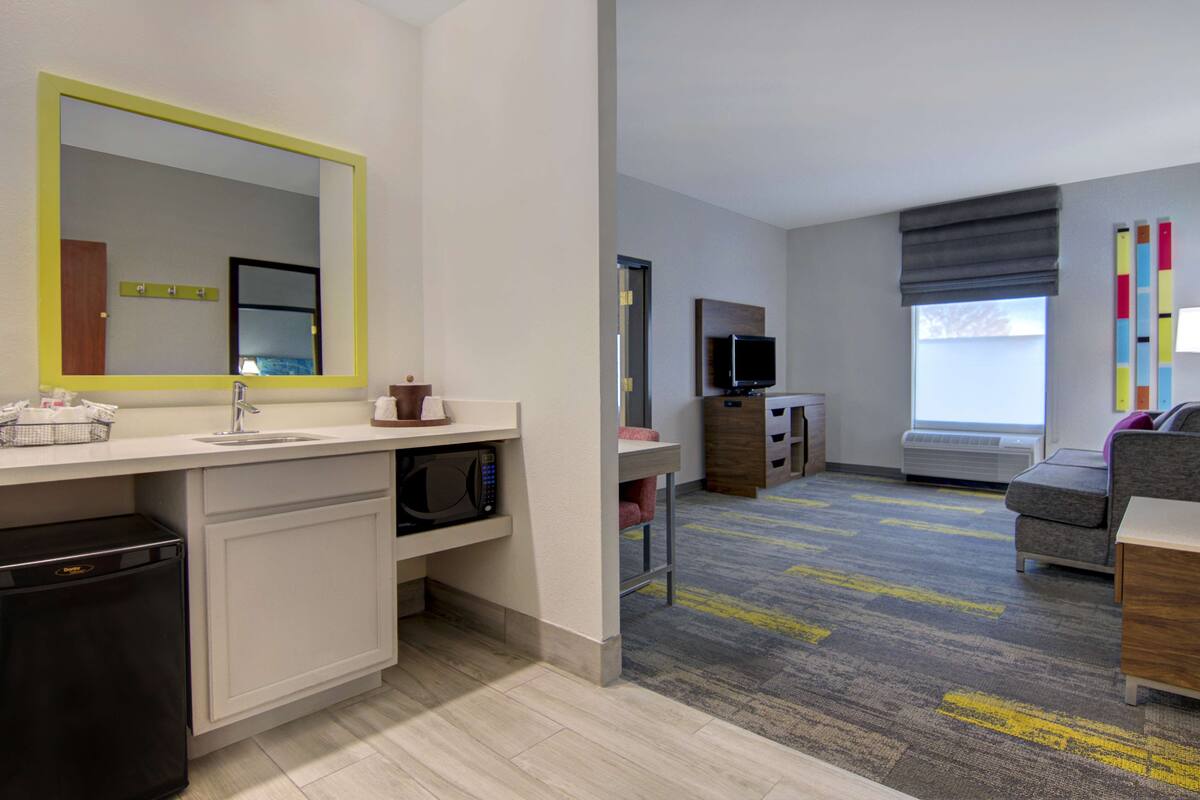 A hotel room with a kitchenette, a large mirror, a flat-screen TV, a sofa, and a window with blinds.