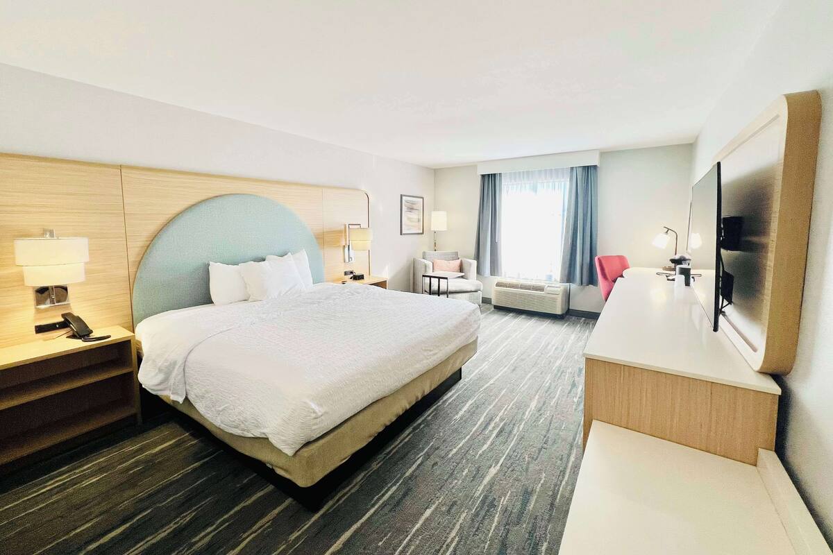 A hotel room with a large bed, bedside tables with lamps, a desk with a chair, a TV, and a window with curtains.