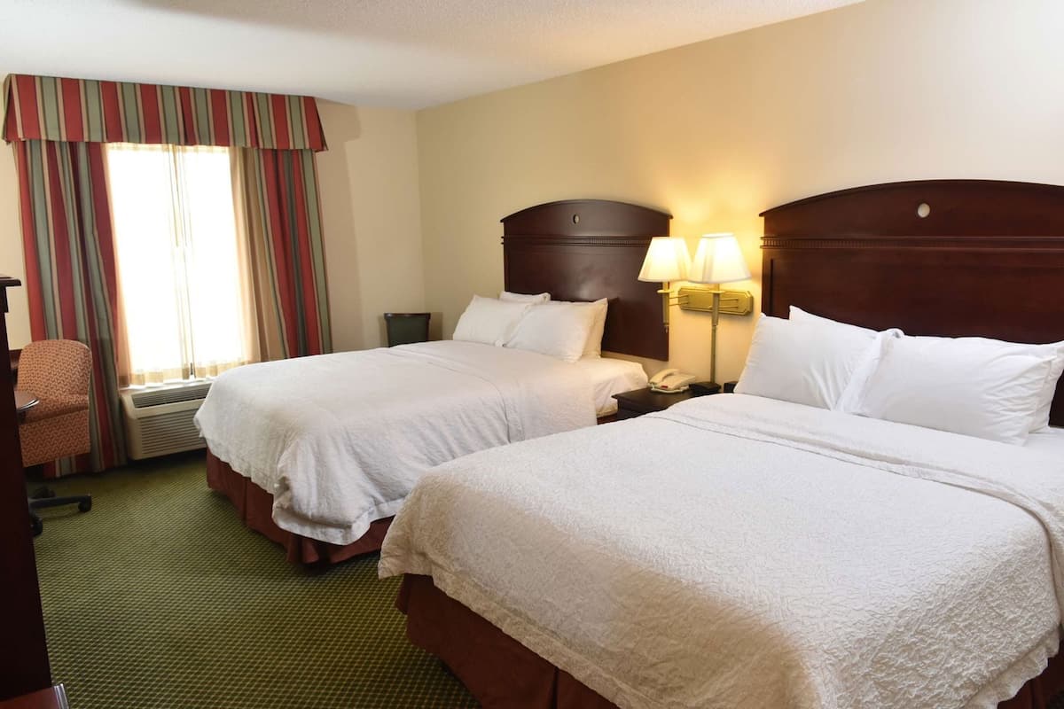 A hotel room with two beds, a desk, a chair, a lamp, and a window with curtains.