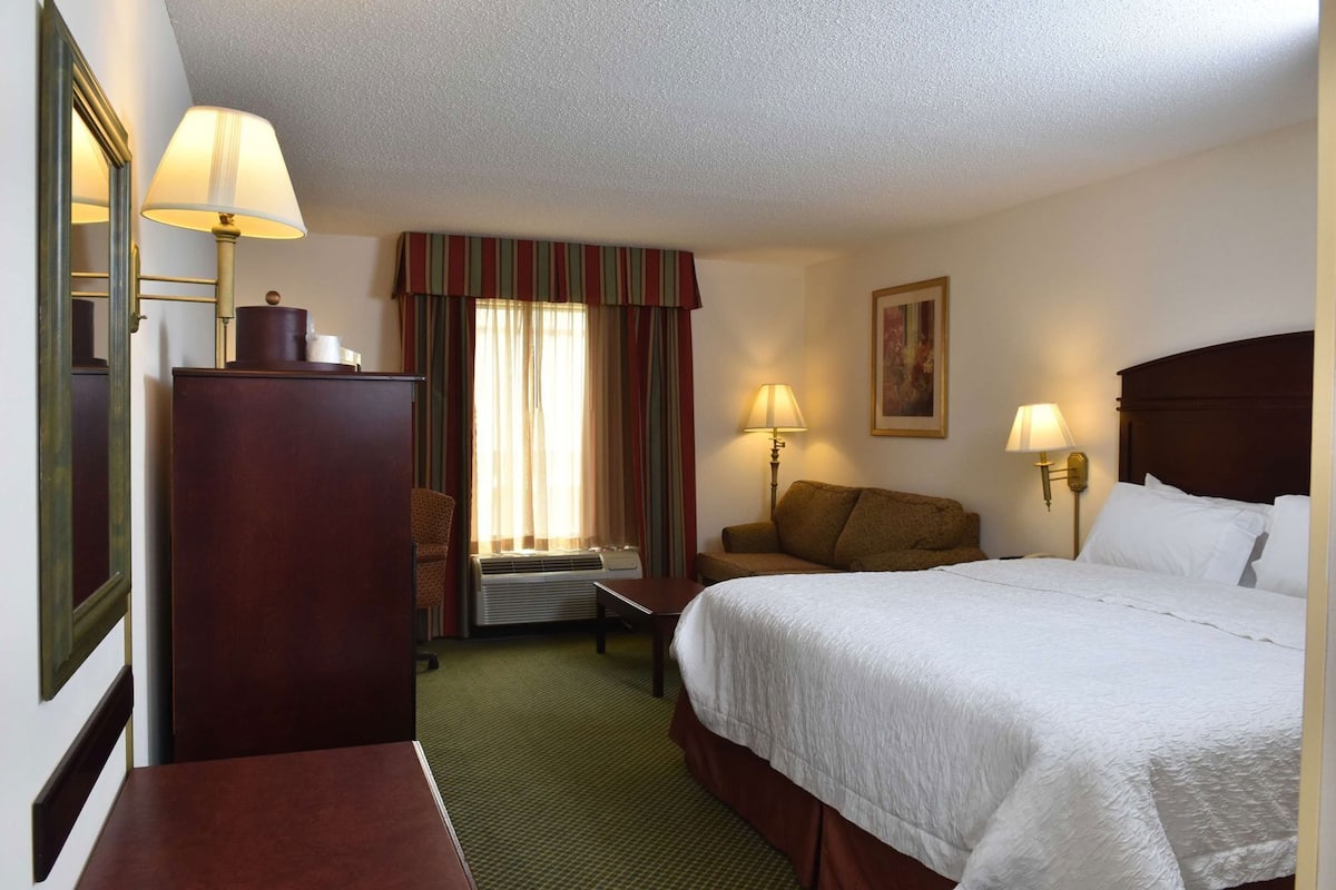 A hotel room with a bed, a desk, a chair, a dresser, a window with curtains, and two wall-mounted lamps.