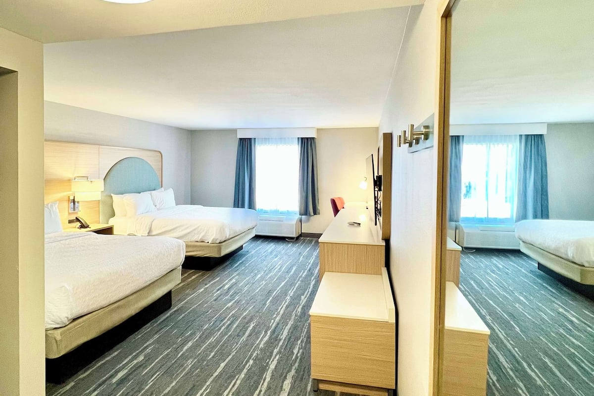 A hotel room with two beds, a desk, a mirror, and a large window with curtains.