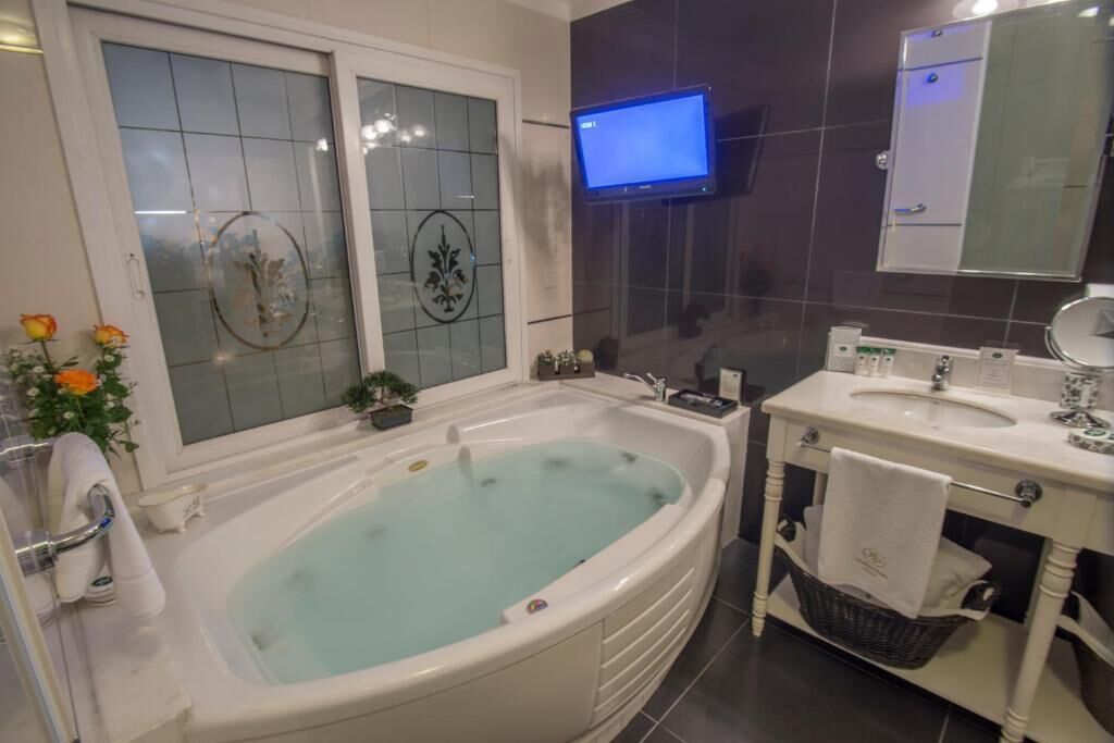 Un baño con bañera, lavabo, televisor montado en la pared y una ventana con paneles de vidrio decorativos.