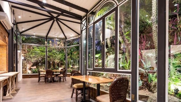 A conservatory with a glass enclosure, wooden tables, and chairs, surrounded by lush tropical plants. - Hotel Villa Morra Suites (Asunción)