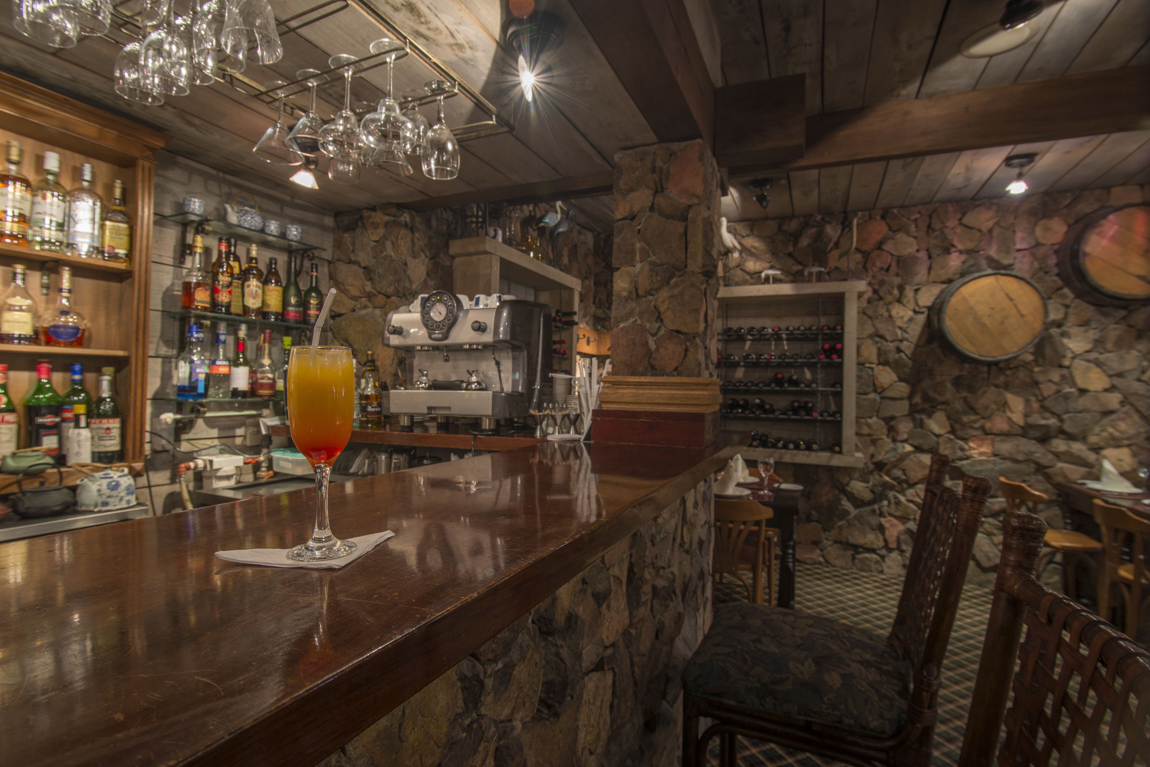 Zona de bar con mostrador de madera, una bebida en un vaso y estantes llenos de botellas de licor.