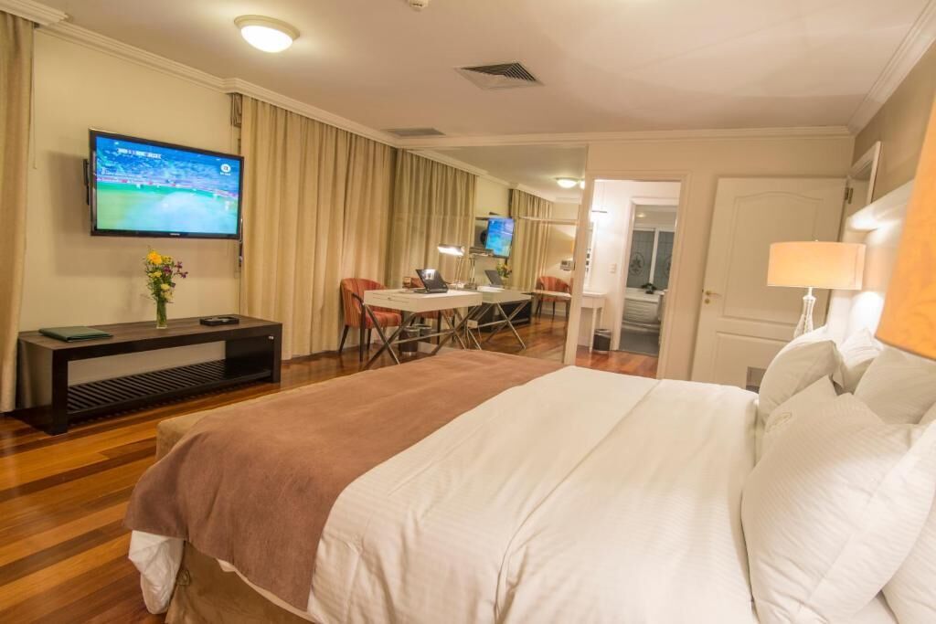 A hotel room with a large bed, a TV on the wall, a desk with a chair, and a dining area.