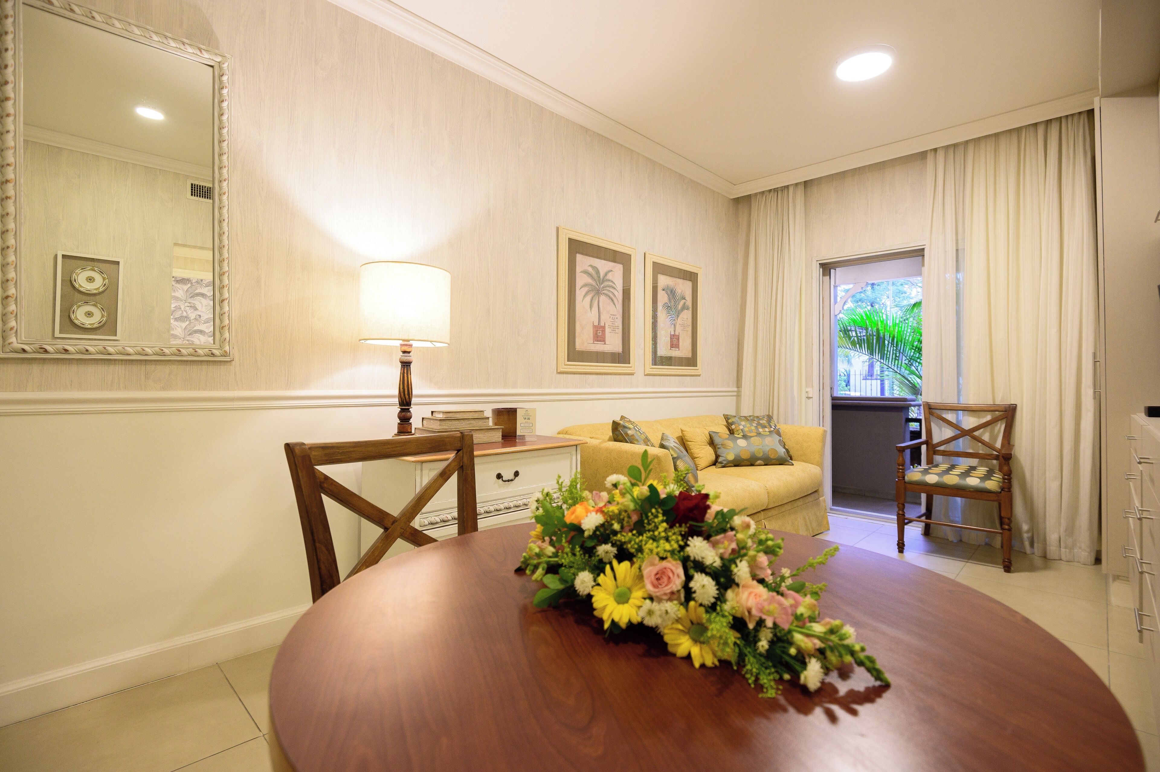 Un comedor con una mesa de madera, sillas, un centro de mesa floral, una lámpara, un sofá y un espejo grande en la pared.