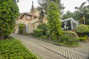 A residential area with a curved driveway, lush greenery, and a house with a brick facade and large windows. - Hotel Villa Morra Suites (Asunción)